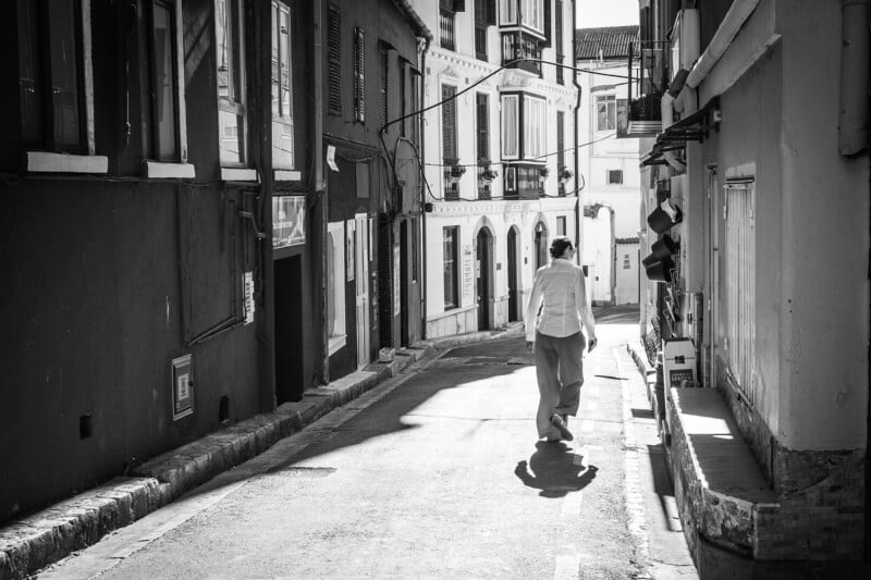 A person walks alone down a narrow, empty street lined with buildings in a sunny, urban setting. The street is partially in shadow, and the person is wearing light-colored clothing. The scene is captured in black and white.