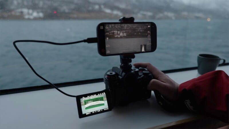 A person in a red jacket operates a digital camera with a smartphone attached on top. Both screens display an outdoor scene with water. The setup sits on a windowsill, and a cup is nearby.