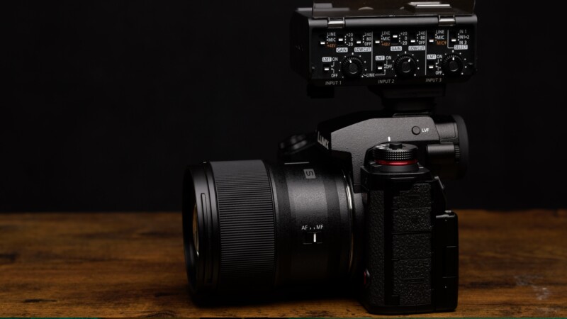 A black DSLR camera fitted with a large lens and an audio input attachment on top sits on a wooden surface against a dark background. The camera features multiple dials and buttons, highlighting its professional design.