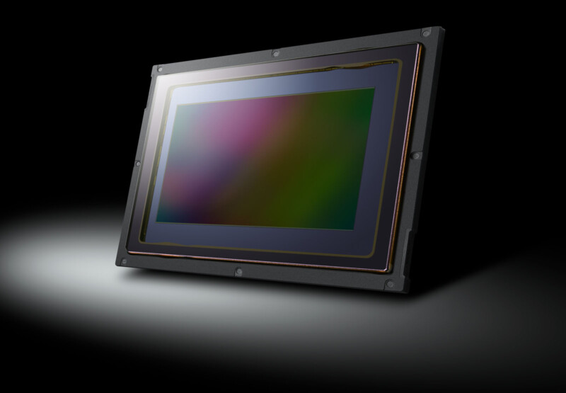 Close-up of a large, rectangular image sensor with a reflective surface, mounted within a black frame. The sensor is angled slightly, set against a dark background with a spotlight effect on one side.