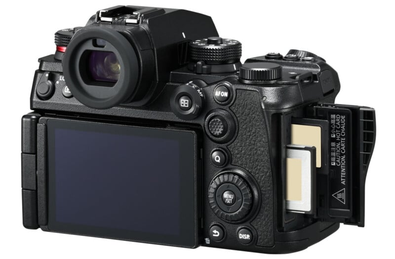 A black digital camera viewed from the back, showing its LCD screen and various control buttons. The side compartment is open, revealing dual memory card slots, each containing a memory card.