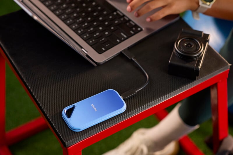 A person using a laptop with an external hard drive connected by a cable. A camera is placed nearby on a black and red table. The person's hand and part of their arm are visible, wearing a watch and bracelet. The background shows green flooring.