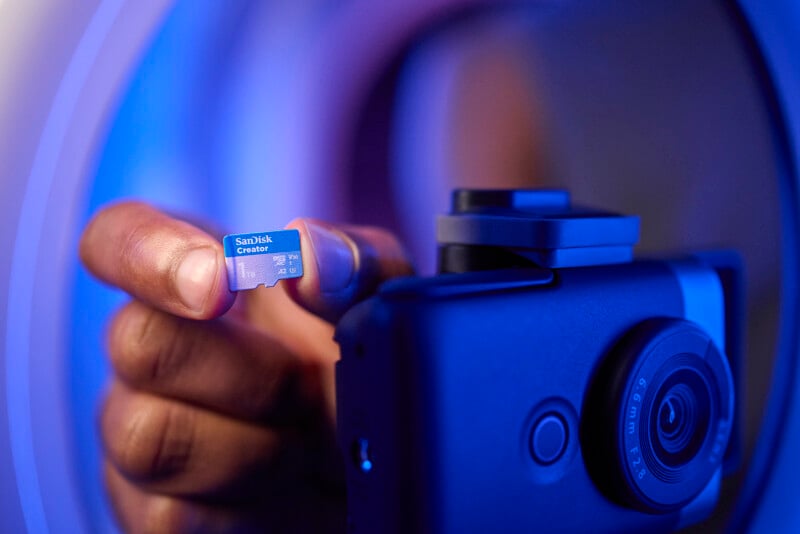 A person holds a SanDisk Creator microSD card near a digital camera. The scene is illuminated with a blue light, creating a futuristic atmosphere. The camera lens is visible in the foreground.