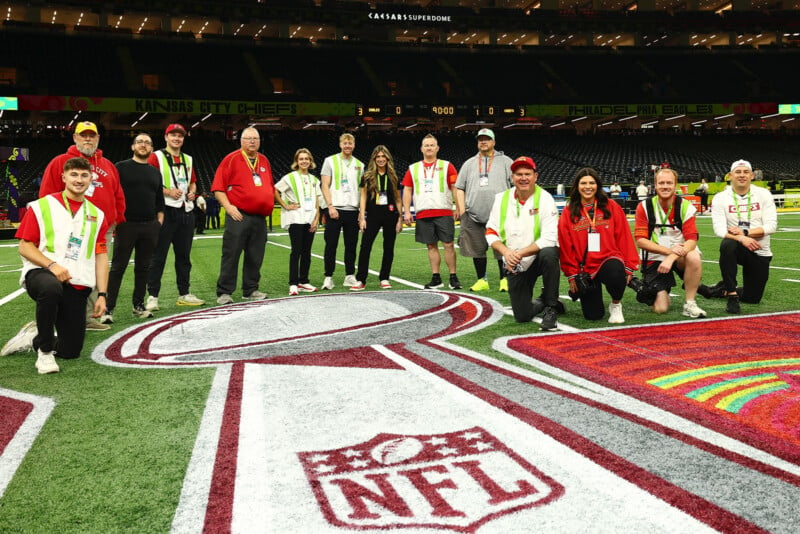 A group of people, some in red and green attire, pose on a football field featuring the NFL logo. They appear to be part of the event staff, gathered inside a large, illuminated stadium.