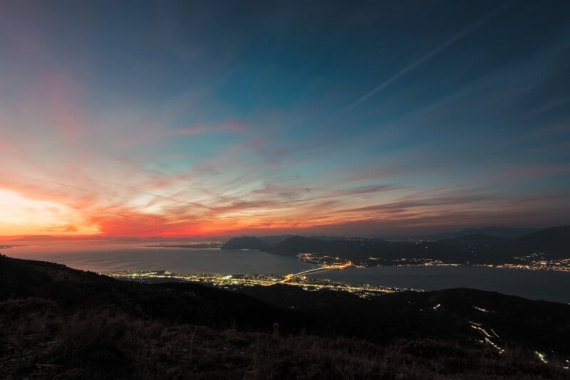 A scenic view from a hillside at twilight, showing a vast landscape with a glowing sunset sky and the sea. City lights illuminate the coastline, with distant mountains silhouetted against the vibrant hues of the sky.