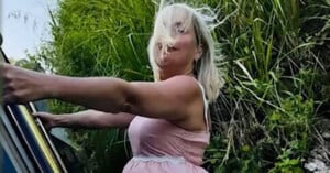 A person with blonde hair in a pink dress is joyfully leaning out of a vehicle, with their hair blown by the wind against a backdrop of lush green foliage.