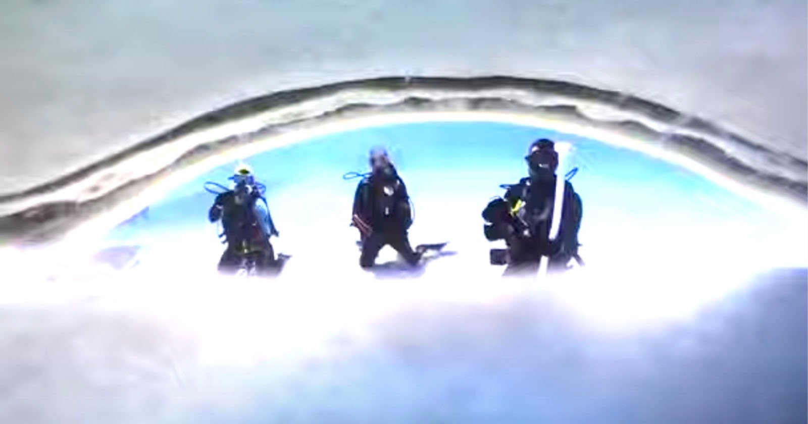 Three scuba divers, seen through a large ice opening, are emerging from an underwater dive. They are in dark wetsuits with tanks and masks, set against a bright, icy backdrop above them.