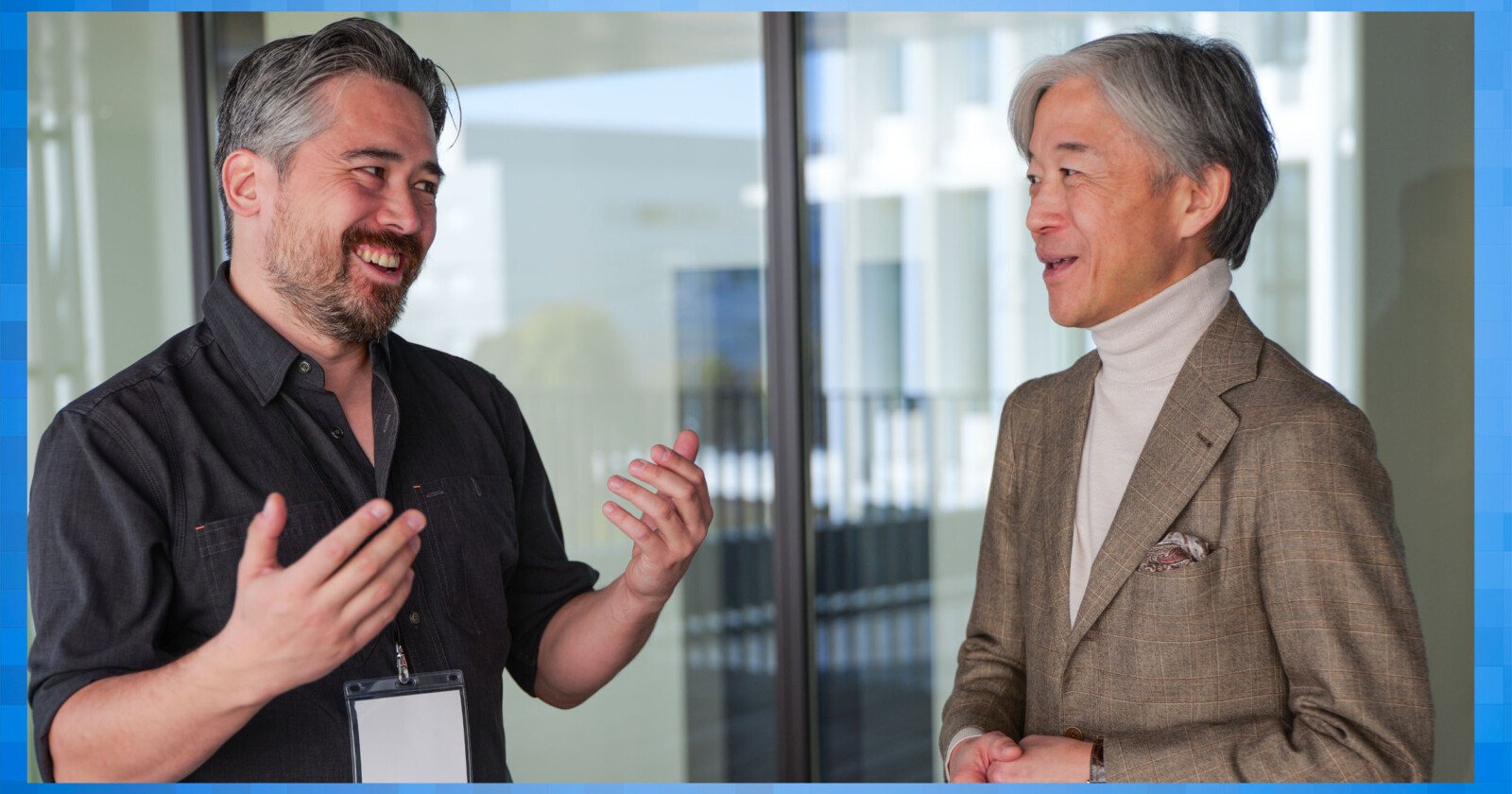 Two men are standing and smiling at each other in a professional setting. One man, with gray hair, wears a black shirt and lanyard. The other, with gray hair, wears a beige jacket and turtleneck. They appear to be engaged in a friendly conversation.