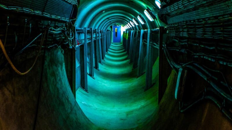 A narrow, curved underground tunnel with greenish-blue lighting. The walls are lined with metal racks and cables, leading to a bright blue door at the end. The ceiling is arched, and the atmosphere is industrial.