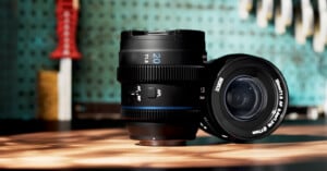 A camera lens with a 20mm focal length is placed upright on a wooden surface, with another lens lying beside it. The background has a blurred, teal pegboard with tools hanging on it. Warm sunlight casts soft shadows over the lenses.