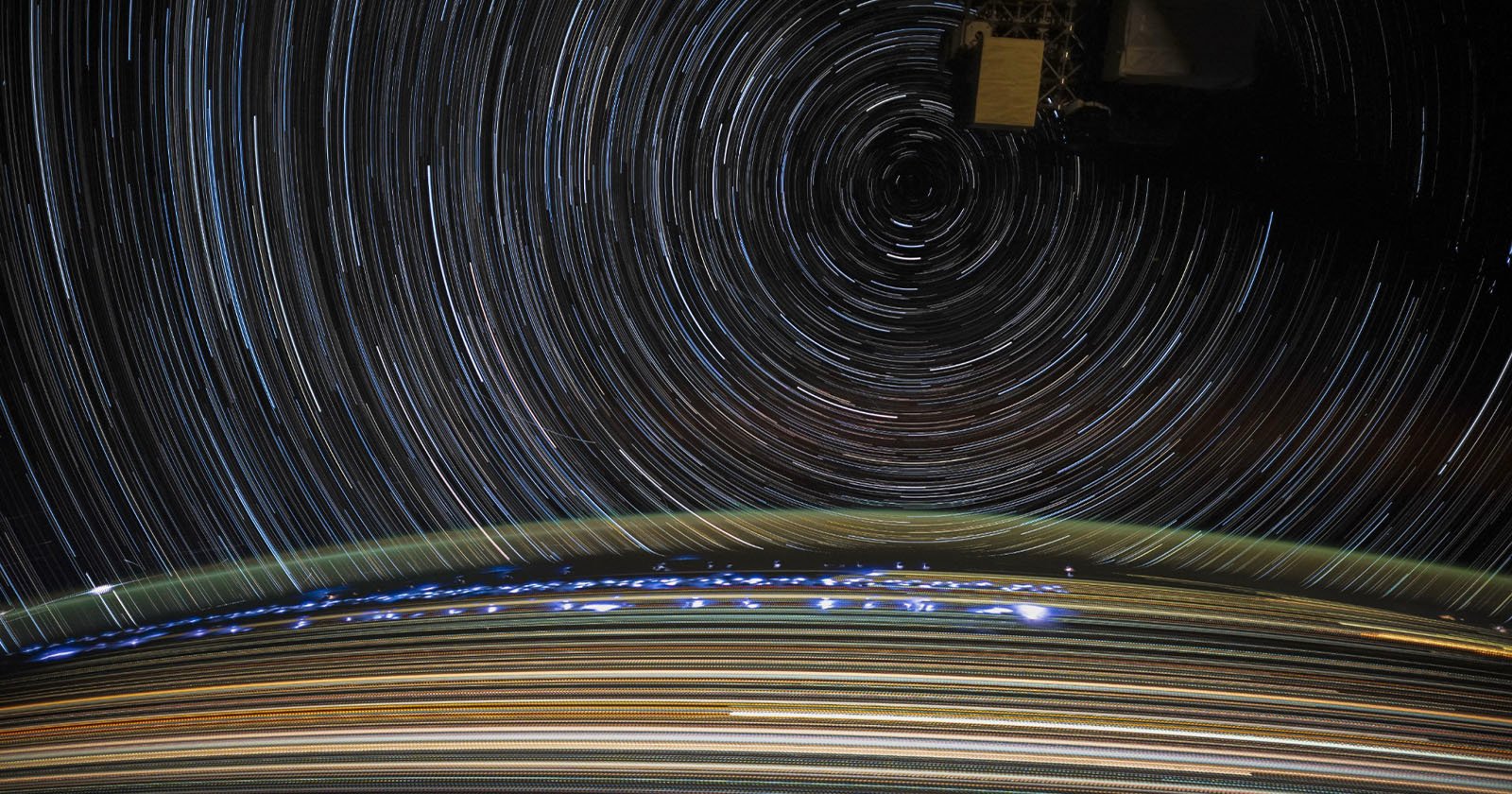 A long-exposure photo from space showing circular star trails in the sky above a glowing Earth. The stars create concentric circles around a central point, while city lights and atmospheric glow are visible on the Earth's surface.