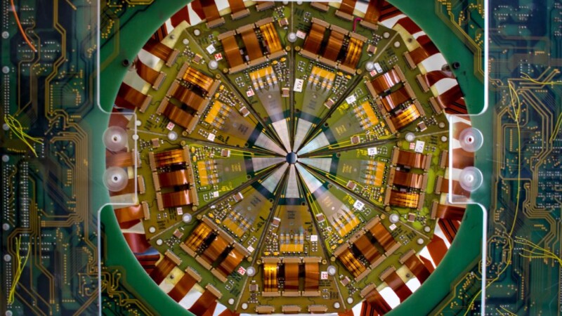 Circular close-up of a complex electronic circuit board with symmetrical radial design. Various components, including resistors and capacitors, are arranged in a starburst pattern, surrounded by a green border with circuit pathways.