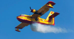 A yellow and red firefighting airplane releases water against a clear blue sky. The aircraft features visible propellers and numbered markings on its body.