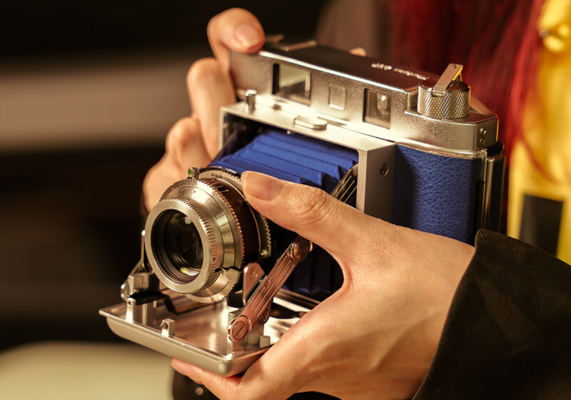 Close-up of a person holding a vintage folding camera with a metallic body and blue bellows. The hands are adjusting the lens, and the background is softly blurred.