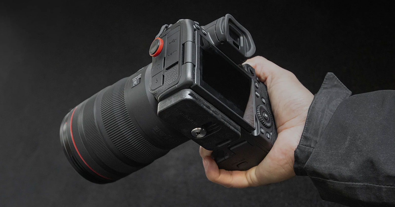 A person holds a professional DSLR camera with a large lens against a dark background. The camera has a flip-out screen and a variety of buttons and dials, with the person's hand gripping it securely.