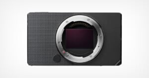 A minimalist black digital camera body with a textured surface, featuring a prominent lens mount in the center. The camera is displayed against a plain light background, showcasing its compact and modern design.