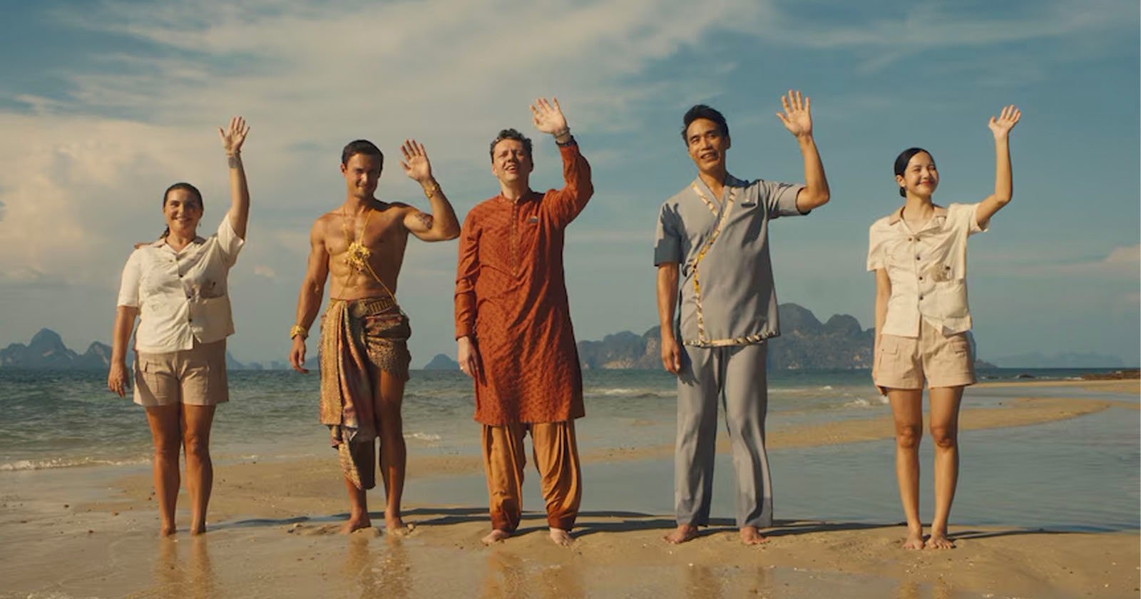 Five people stand on a beach, smiling and waving. They wear diverse outfits: a uniform, traditional attire, a bright orange outfit, casual blue attire, and another uniform, reflecting a mixture of cultural styles. The sea and sky are in the background.