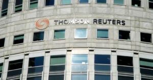 A curved building facade featuring the Thomson Reuters logo and name. The logo consists of an orange circular design. Multiple windows are visible on the stone-clad structure.