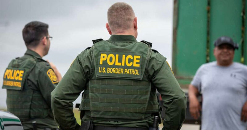 Two U.S. Border Patrol officers in green uniforms face away, wearing vests with "POLICE U.S. BORDER PATROL" in yellow. They stand near a man in a gray shirt and cap, with a green truck visible in the background.