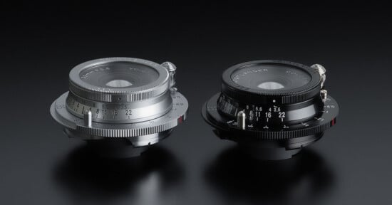 Two camera lenses are displayed side by side on a reflective black surface. The lens on the left is silver with engraving, while the lens on the right is black with similar features. Both lenses have textured grips and marked aperture settings.