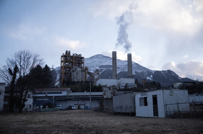 Industrial facility with tall chimneys emitting smoke, set against a snowy mountain and cloudy sky. Foreground features bare trees and trailers, suggesting a cold, rural environment.