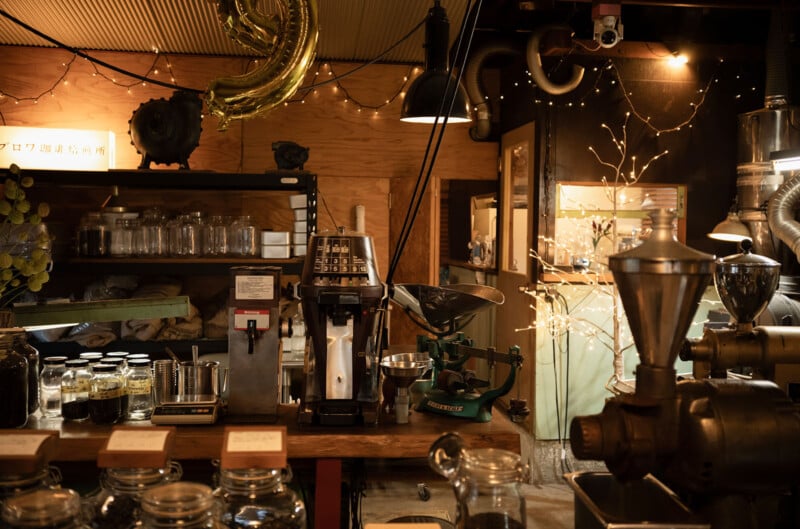 Cozy coffee shop interior with wooden walls, featuring jars on shelves, a coffee grinder, and a weighing scale on a counter. Warm lighting creates a welcoming atmosphere, enhanced by decorative string lights and a gold balloon.