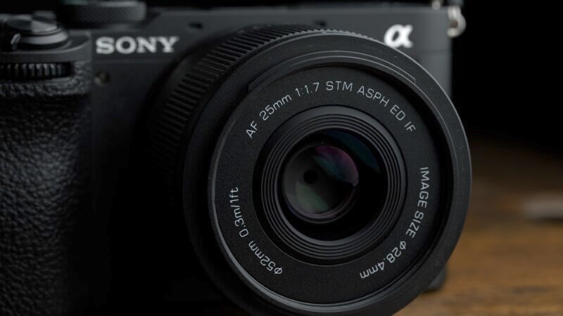 Close-up of a black camera lens attached to a camera body, featuring text indicating it's a 25mm f/1.7 lens. The camera body has a textured grip and bears a logo with a stylized "a" symbol. The background is dark and blurred.