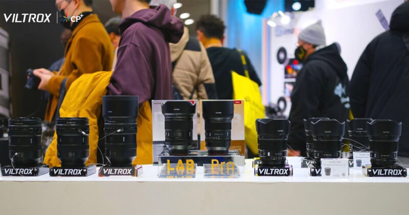 A group of people in a booth with various Viltrox camera lenses displayed on a table. Some individuals are wearing jackets and face masks. Bright lighting and a busy atmosphere suggest an exhibition or trade show setting.