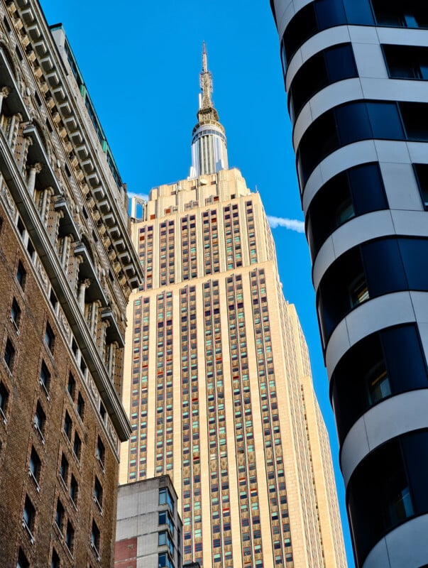 View of a tall building with a spire, seen from below, framed by two other buildings on either side. The sky is clear and blue, and the architecture features a mix of classic and modern styles.