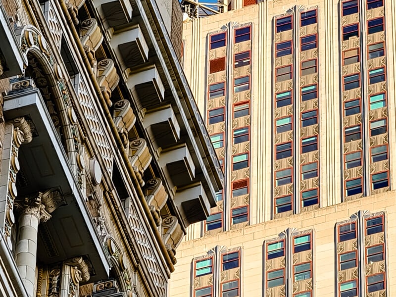 Close-up of two buildings; one with ornate, classical columns and detailed cornices on the left, and a tall, beige building with vertical lines and repetitive window patterns on the right. Both display architectural contrast against a clear blue sky.