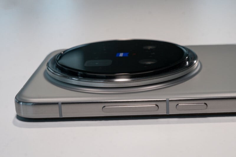 Close-up of a smartphone lying on its side, showcasing a prominent circular camera module on the back. The phone has a metallic frame with visible buttons.