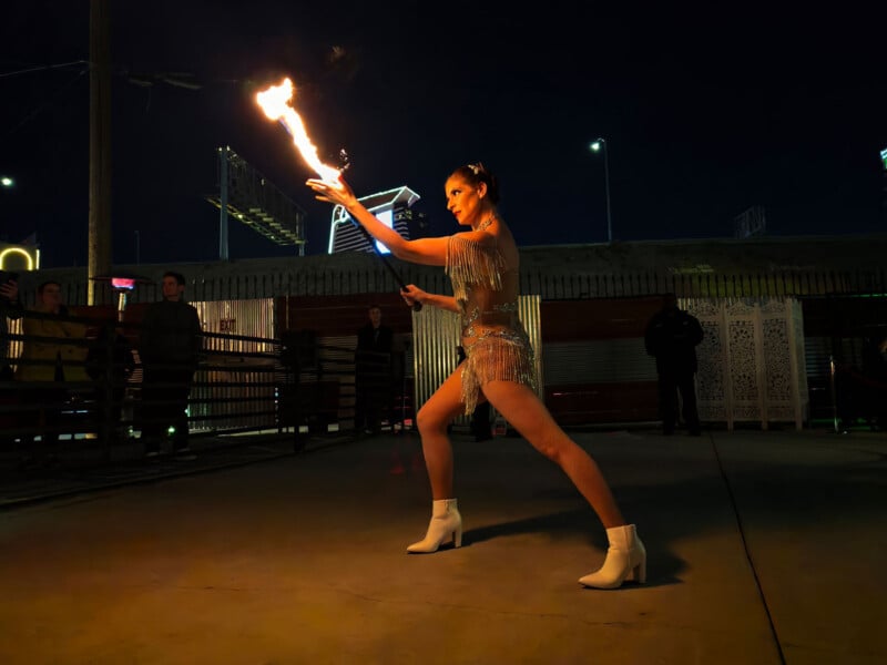 A performer in a sequined outfit and white boots skillfully juggles fire at night. The flame illuminates the scene against a dark backdrop, with onlookers in the background.