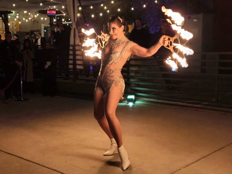 A performer in a sparkling outfit skillfully spins flaming poi at an outdoor event. The scene is lit by overhead string lights, and an audience watches in the background, captivated by the fiery display.