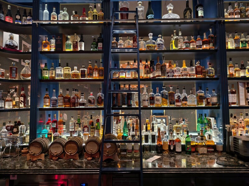 Dimly lit bar with shelves filled with various liquor bottles and a few mini wine barrels. A ladder leans against the shelves for access. The countertop below is cluttered with bottles and glassware, creating an inviting atmosphere.