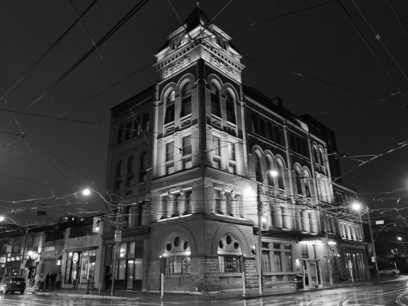 A black and white photo of a historic, multi-story brick building on a city street corner at night. The building is lit by spotlights, highlighting its arched windows and decorative details. Streetlights and overhead wires are visible.