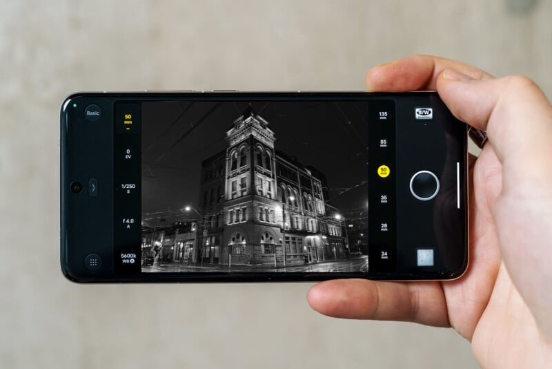 A person holding a smartphone horizontally, displaying a black-and-white photograph of a historic multi-story building at night. The building is lit by streetlights, and photography settings are visible on the phone screen.