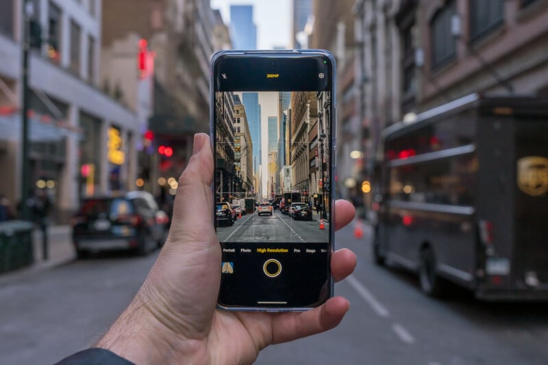 A hand holds a smartphone in the foreground, capturing a busy urban street scene. The phone's screen displays the same street view, showing buildings, cars, and a bright sky. The background depicts a bustling city street with parked vehicles.