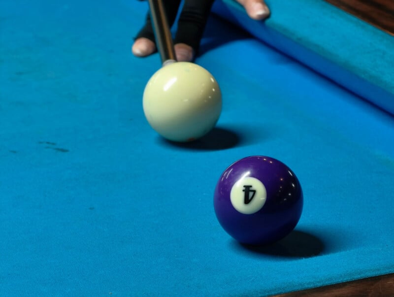 A close-up of a person playing billiards. A cue stick is positioned to hit a white cue ball towards a blue billiard ball labeled with the number four, on a blue pool table.