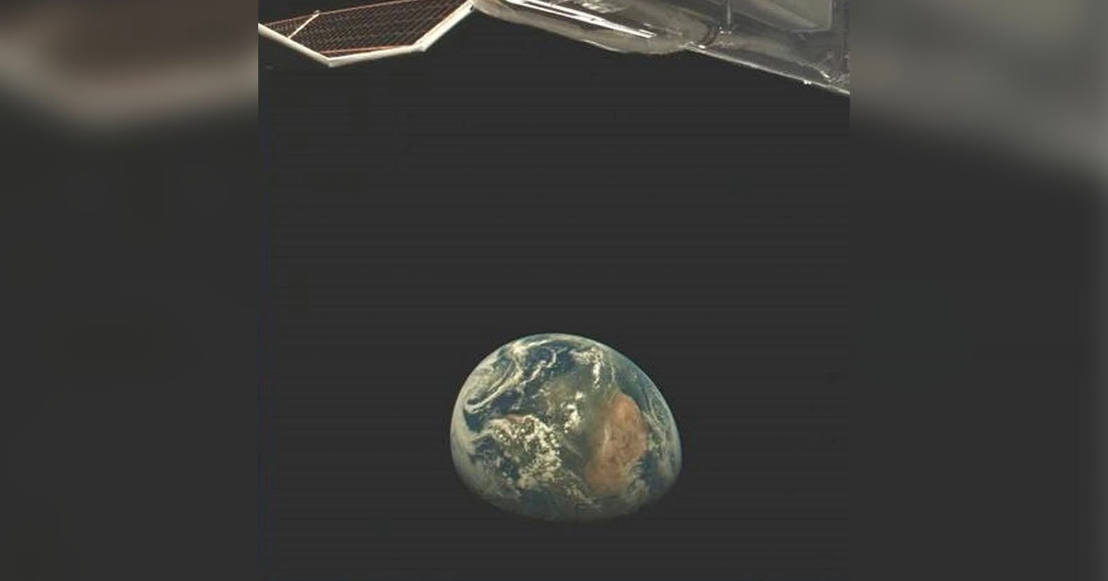 A view of Earth from space, showing a portion of a spacecraft or satellite panel in the upper left corner. The planet appears small against the vastness of space, with visible cloud formations and parts of the continents.