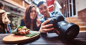 A smiling couple sits at an outdoor table sharing food wrapped in banana leaves, next to a wooden platter. One person holds a camera, showing the display to the other, capturing a moment together in a street market setting.