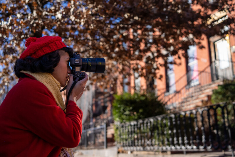 A person wearing a red coat and hat uses a camera with a long lens to photograph a row of brick buildings. The scene is set outdoors with sunlight filtering through bare tree branches in the background.