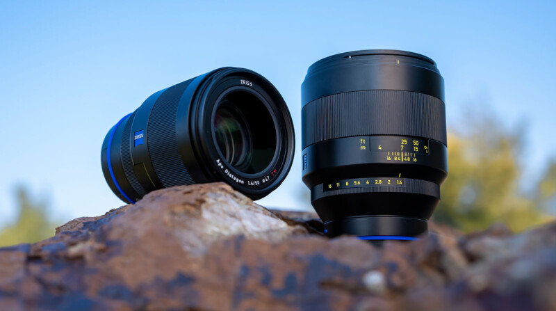 Two camera lenses with black bodies are positioned on a rocky surface outdoors, with a blurred background of trees and a blue sky. The lens on the left is slightly tilted, showcasing its glass front.