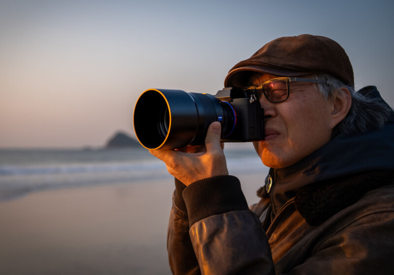 A person wearing a brown cap and glasses is holding a camera up to their face, taking a photo at a beach during sunset. The scene is calm with soft orange lighting and the ocean in the background.