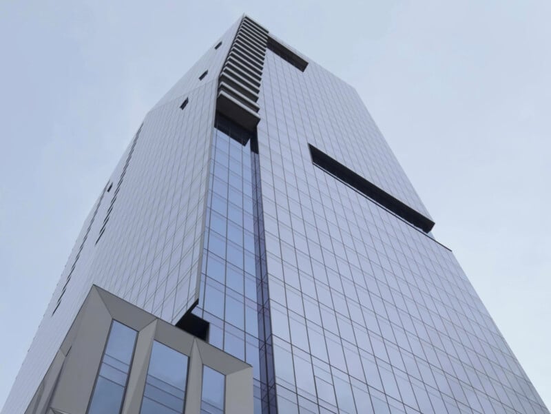 The image shows a tall, modern skyscraper with a sleek glass facade and geometric design elements against a pale blue sky. The building features a series of vertical windows and sections protruding at different angles.