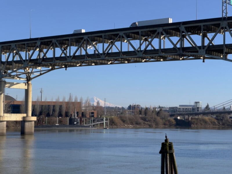 A long bridge spans across a calm river, with vehicles traveling over it. In the background, a distant snow-capped mountain is visible under a clear blue sky. Trees and industrial structures line the riverbank.