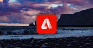 A logo with a red background and a stylized white letter "A" in the center overlays a scenic ocean view. Waves crash on a pebble beach under a dramatic, cloudy sky. Rocky cliffs are silhouetted in the background.