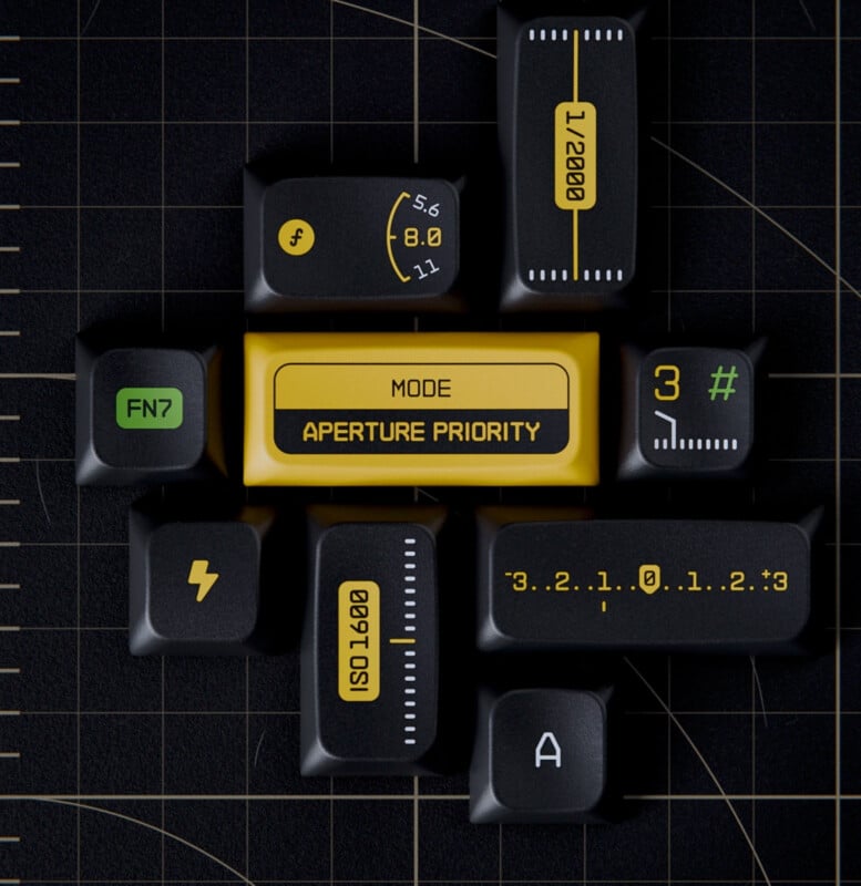 A flat-lay photo of black keyboard keys with photography-related symbols, like ISO, aperture, and shutter speed, arranged around a central yellow key labeled "Mode Aperture Priority." The keys are placed on a black grid surface.