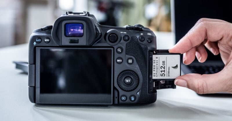 A hand is inserting a 512 GB memory card into the slot of a digital camera placed on a table. The camera's LCD screen and control buttons are visible. In the background, there's a partially visible laptop.