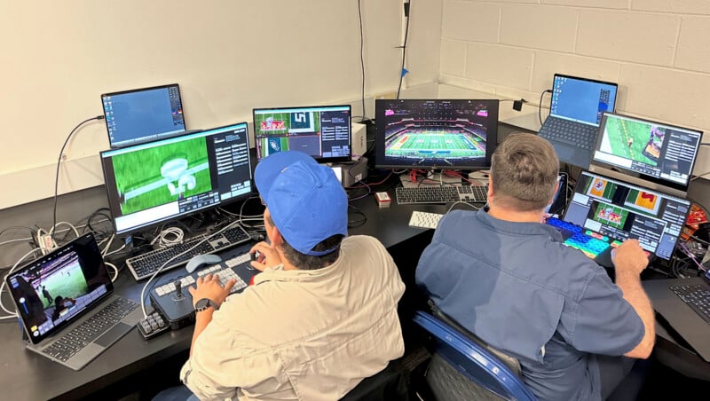 Two people sit in front of multiple computer monitors displaying sports graphics and footage. They appear to be working on video production or editing in an office setting. Both are focused on their tasks, surrounded by cables and equipment.