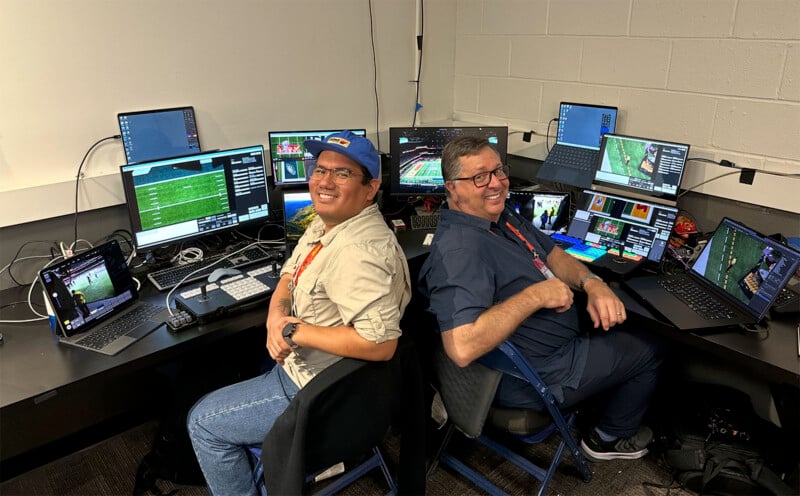 Two people sitting back-to-back at desks filled with several monitors and laptops displaying sports footage. The room has a cluttered yet organized setup, with wires and equipment visible. Both individuals are smiling at the camera.